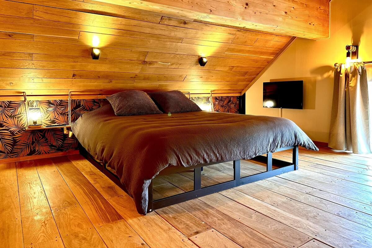 Quarto com teto de madeira, cama com estrutura metálica, televisão fixada na parede e janela com cortinas.