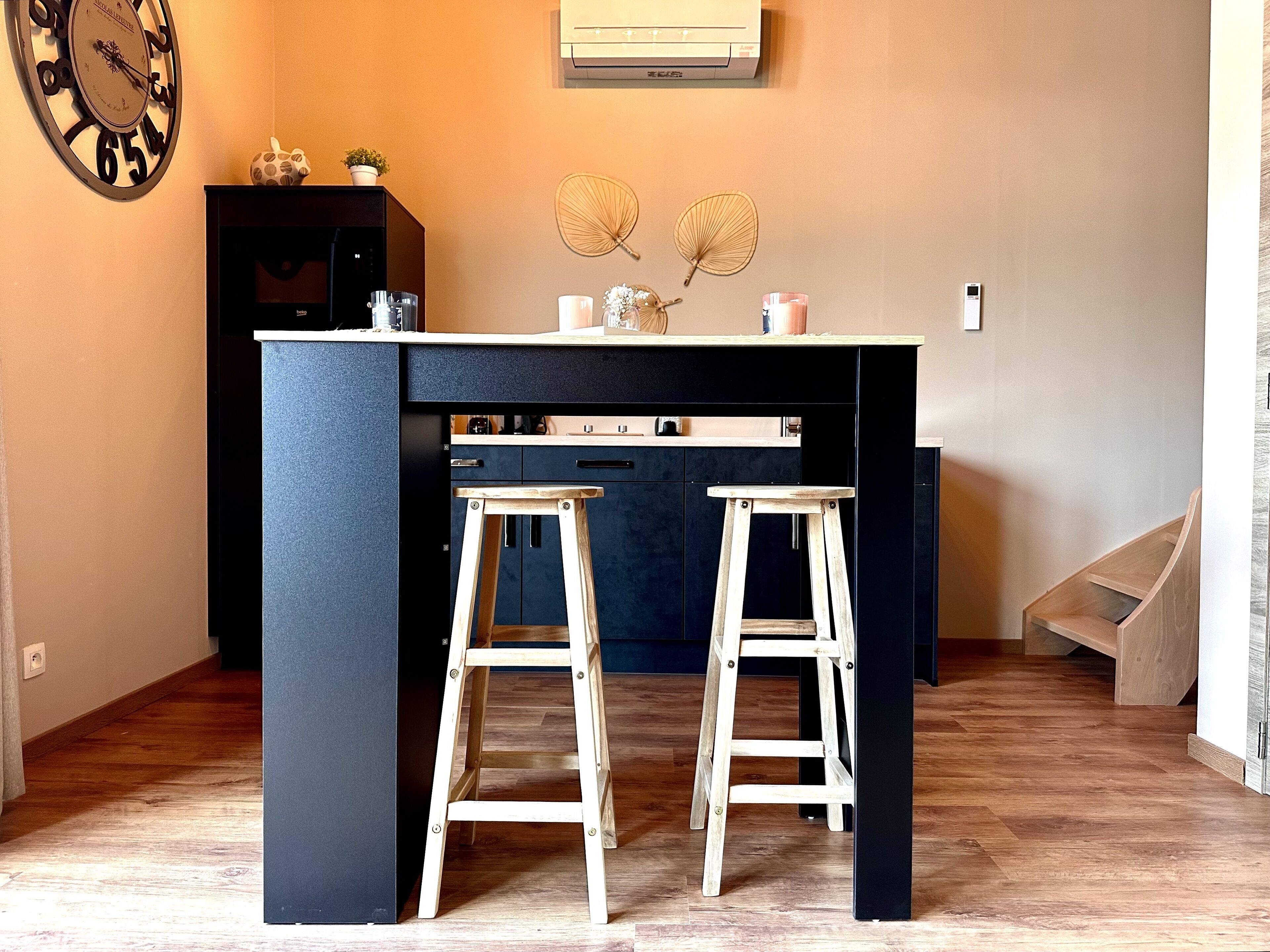 A compact kitchen with a dark blue island, two wooden stools, a clock, and decorative fans on the wall.