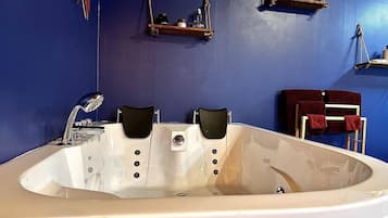 Una bañera grande con dos sillas negras, un grifo blanco y un estante de madera con botellas y un gancho.