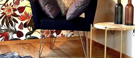 A cozy room with a dark blue armchair, a small round table with two bottles, a fluffy rug, and a vibrant floral wallpaper.