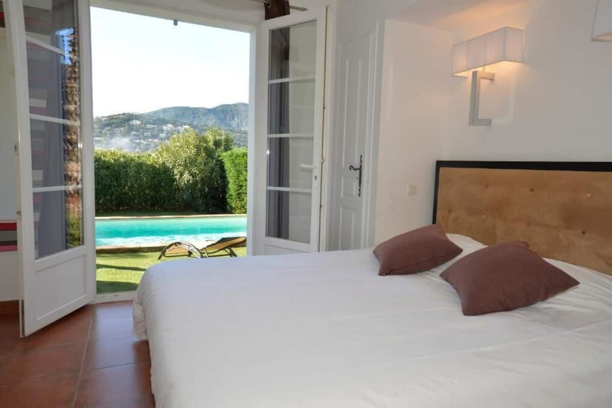 Ein Schlafzimmer mit Bett, zwei Kissen, Blick auf einen Pool und einer Tür zu einem Außenbereich.