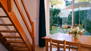 Eine Holztreppe führt zu einem Essbereich mit einem Tisch für zwei Personen, einer weißen Blumenvase und Blick auf einen Garten mit Tisch und Stühlen.