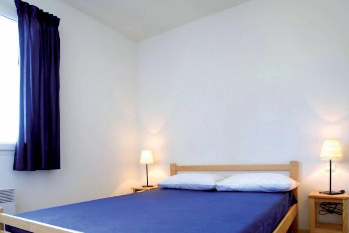 Habitación individual con cama de madera, ropa de cama azul, dos mesitas de noche con lámparas y una ventana con cortinas oscuras.