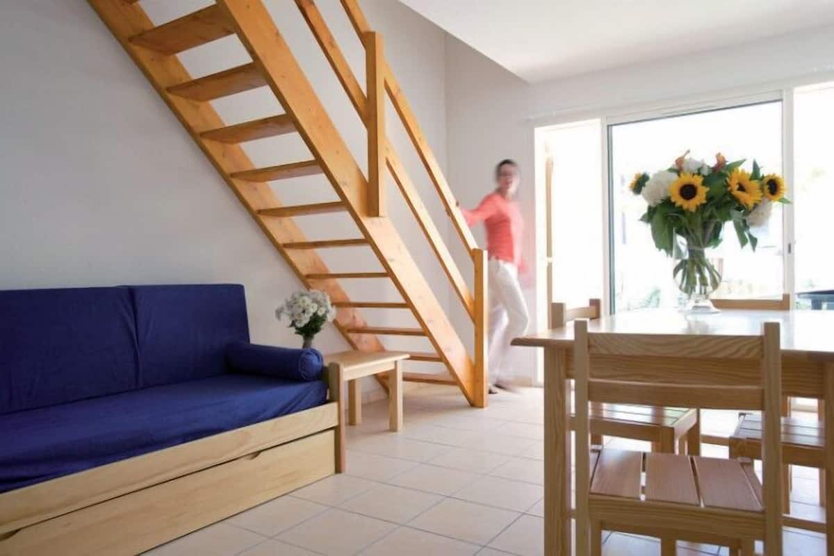 Una sala de estar moderna con un sofá azul, una mesa de comedor de madera y sillas. Se ve una escalera de madera al fondo.
