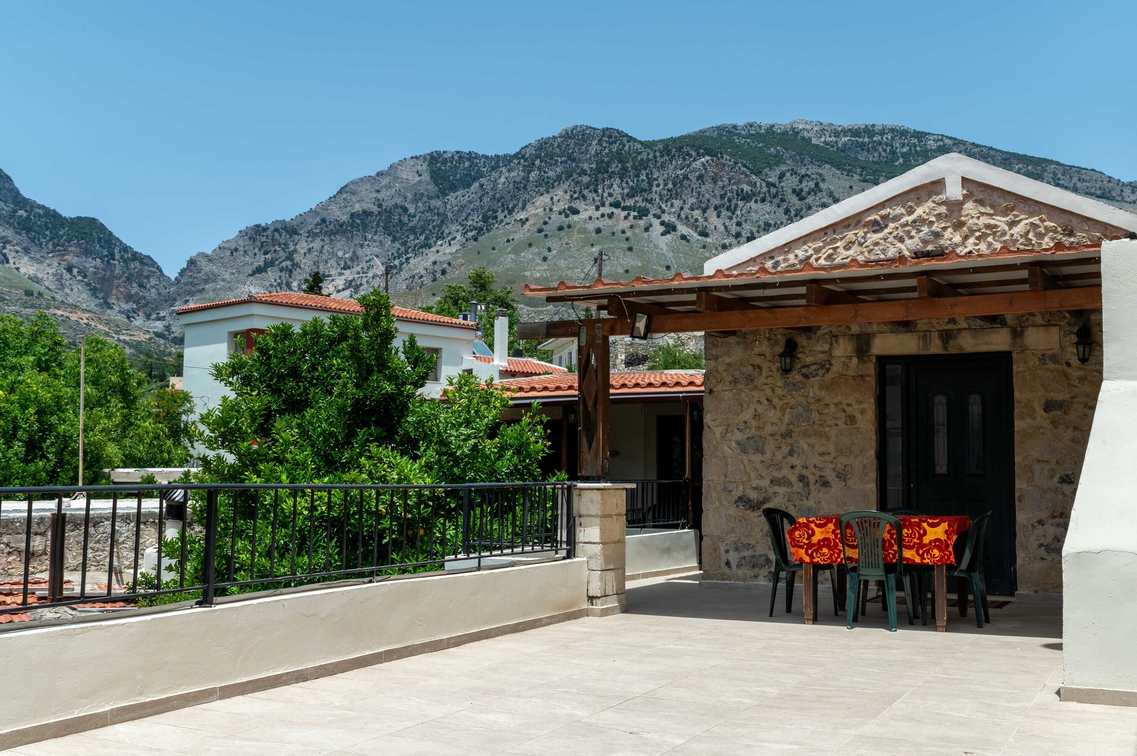 A stone building with a tiled roof, a balcony with a table and chairs, and a mountain backdrop.