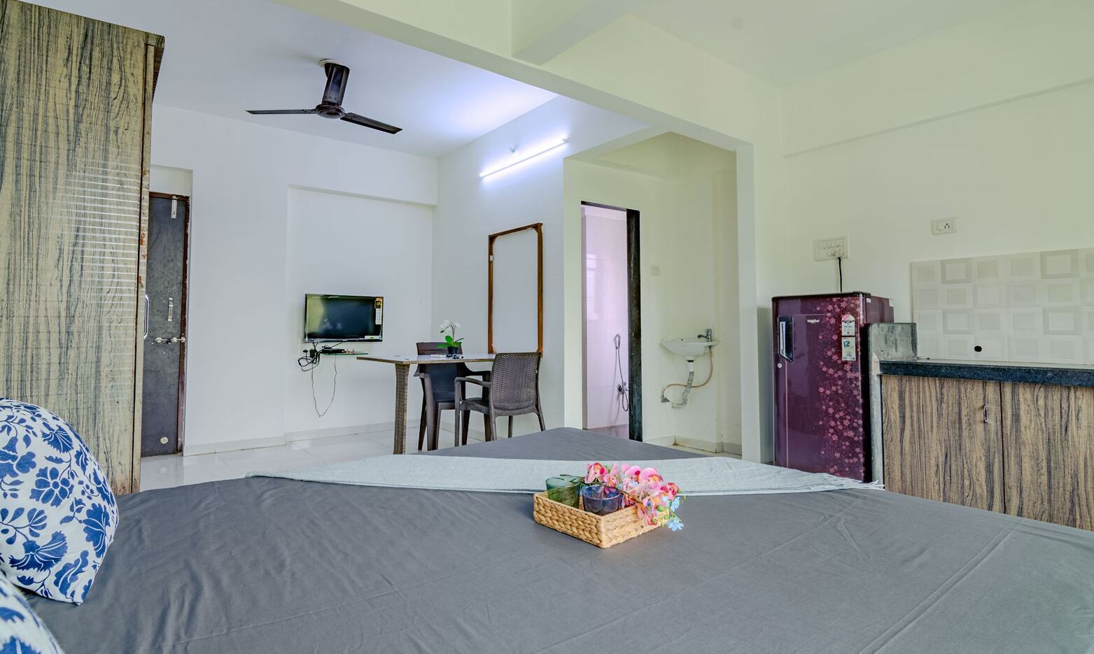 Hotelkamer met een bed, een bureau met stoel, een televisie, een plafondventilator, een koelkast en een badkamer met wastafel en douche.