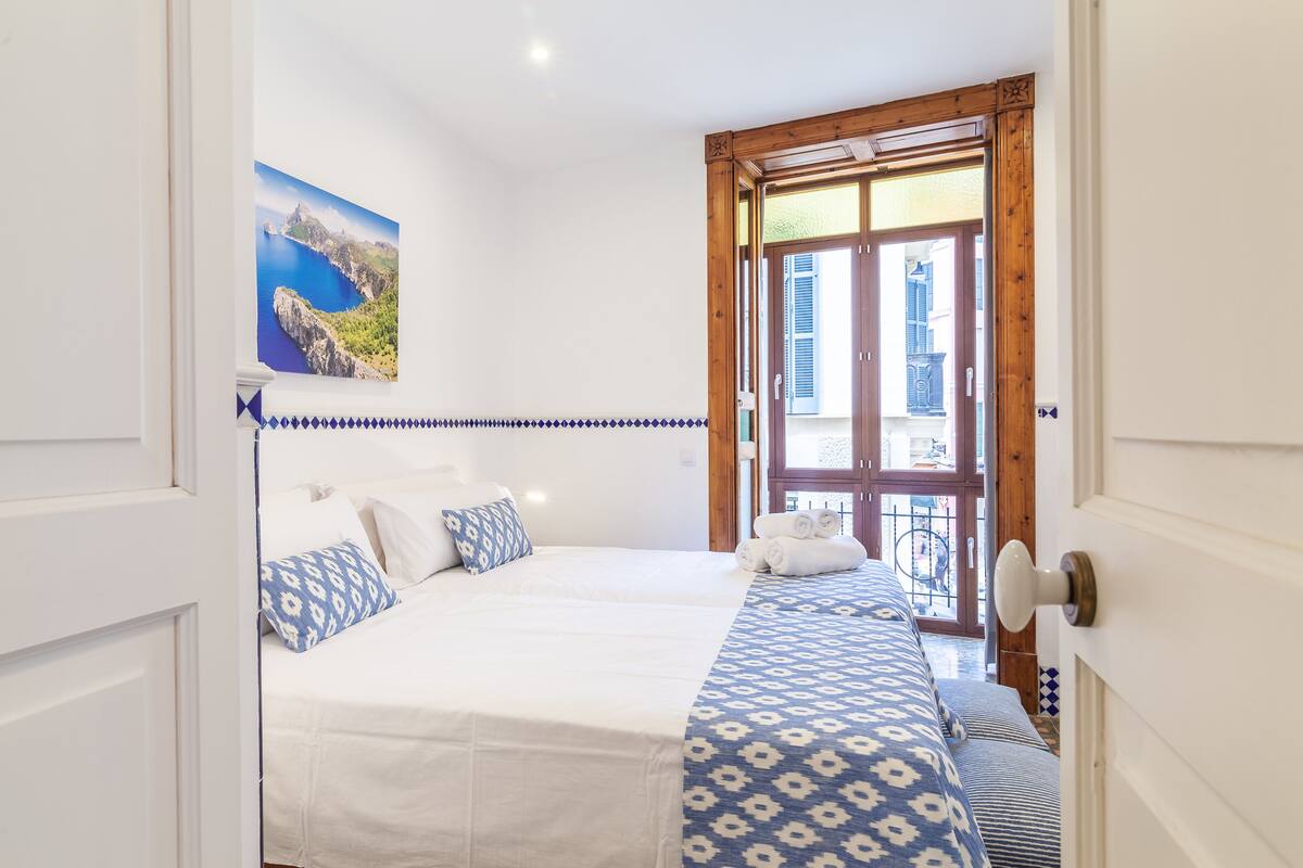 Un dormitorio con cama, almohadas, colcha estampada, un cuadro de un paisaje costero y vistas a través de una puerta de madera.