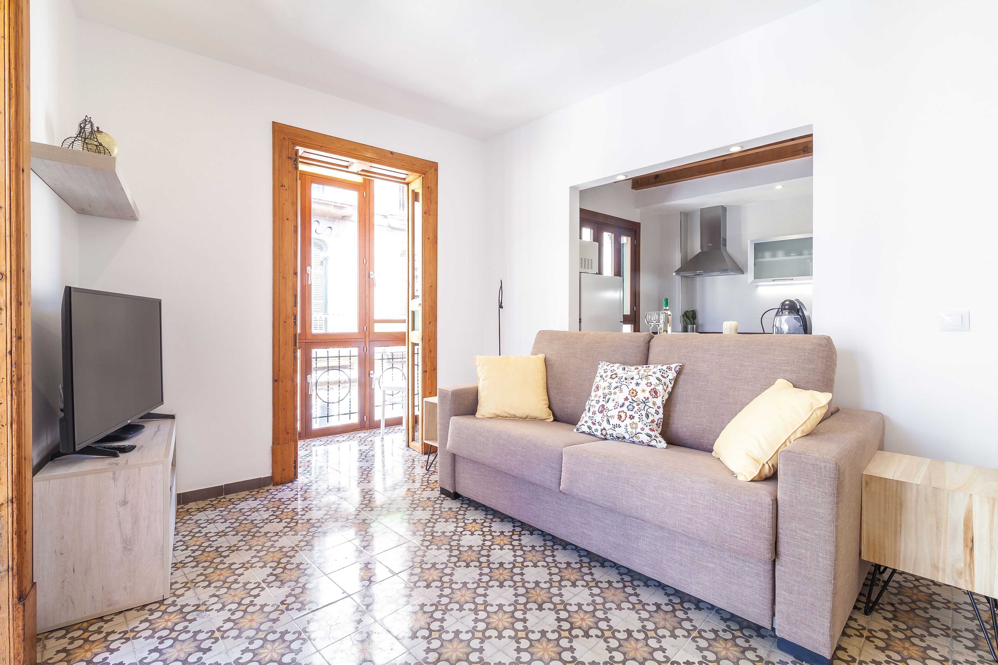 Una sala de estar con un sofá, un televisor sobre un mueble bajo, un suelo estampado y una cocina visible a través de una puerta abierta.