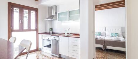 Cocina moderna con armarios blancos, extractor de acero inoxidable y lavavajillas. Cuenta con un comedor con mesa y sillas, y un dormitorio con cama y ventana.