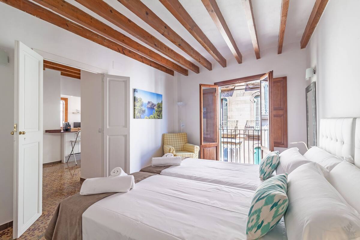 Habitación de hotel con dos camas, un balcón con vistas y un comedor visible a través de una puerta abierta.