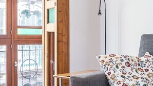Una habitación con un sofá gris, un cojín floral, una lámpara de pie alta y un ventanal con vistas a edificios.