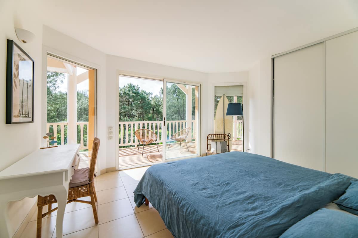 A bedroom with a large bed, a desk, and a chair, with a balcony and outdoor furniture visible.