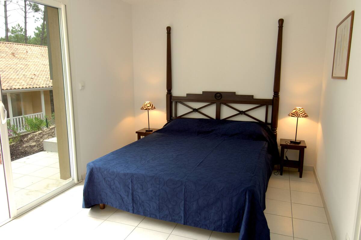A bedroom with a large bed, two bedside tables with lamps, a view through a sliding glass door to an outdoor area, and a framed picture on the wall.