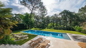 Une piscine avec des chaises longues, un espace patio et un décor boisé en arrière-plan.