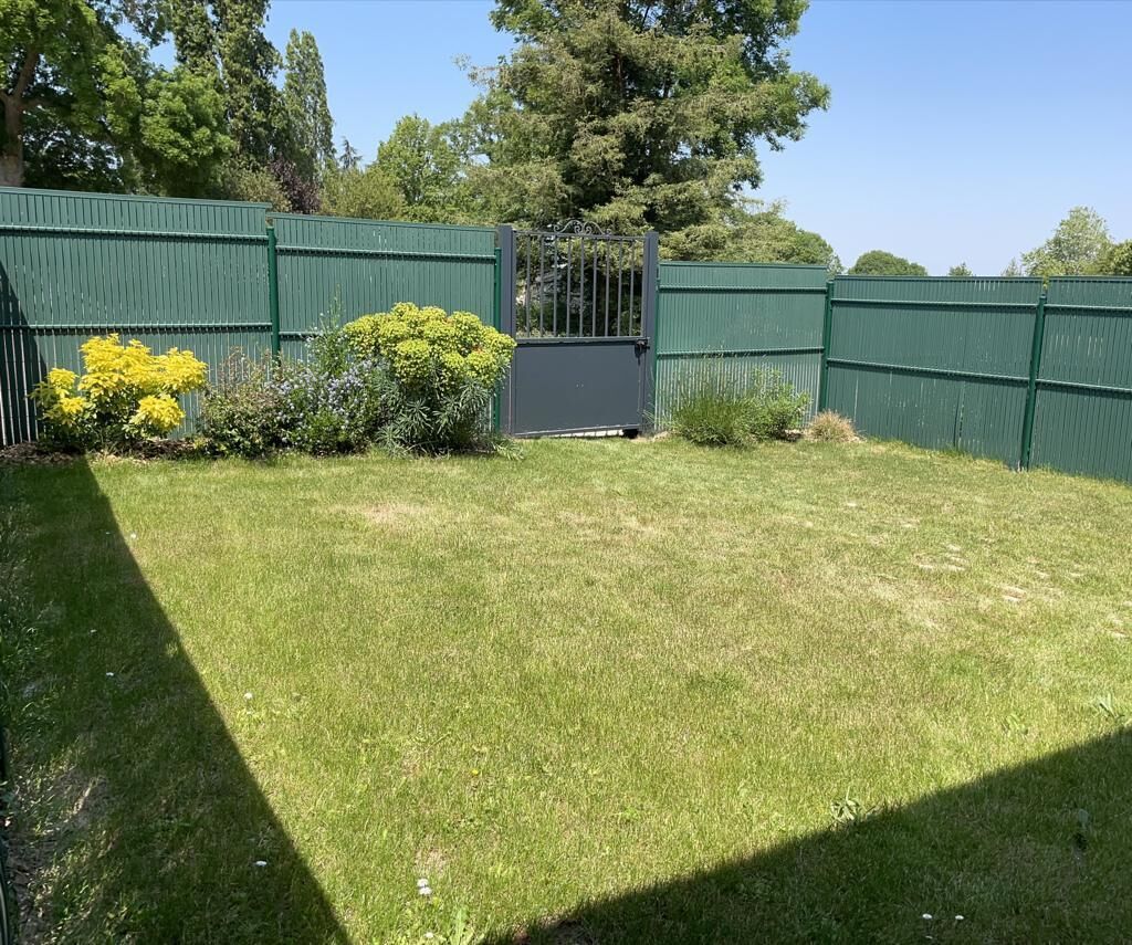 Patio trasero cercado con césped, una puerta y algunos arbustos.