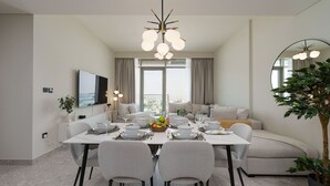 A modern dining area with a white table set for six, surrounded by grey chairs. A chandelier hangs above, and a large mirror is on the right wall.