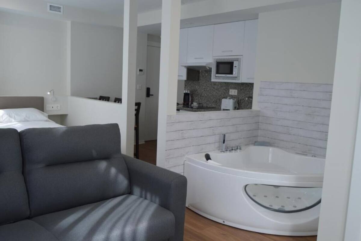 A modern hotel room with a gray sofa, a white bathtub, and a kitchenette with a microwave.