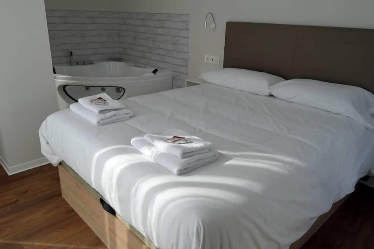 A hotel room with a bed, white linens, a wooden headboard, a white brick wall, and a small white bathtub.