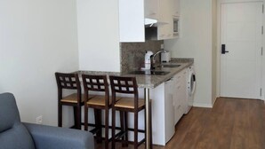 Una cocina compacta con lavadora incorporada, un pequeño comedor con dos sillas de madera y un sofá gris.
