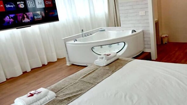 Habitación de hotel con una cama grande, una bañera blanca, un televisor de pantalla plana y un ventanal con cortinas.