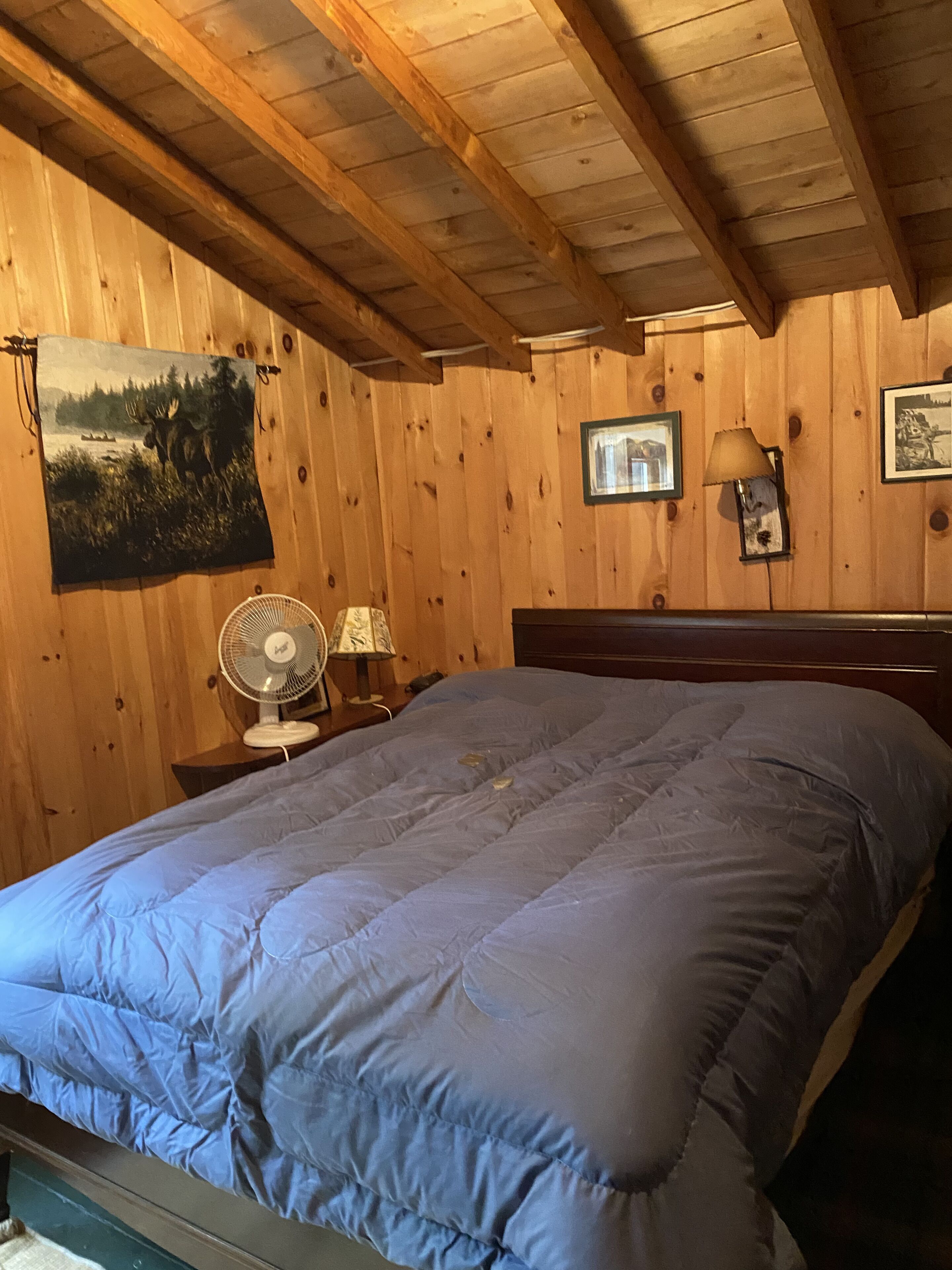 A wooden cabin room with a bed, a fan, a lamp, and a painting on the wall.