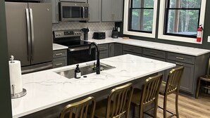 A modern kitchen with grey cabinets, a stainless steel refrigerator, a microwave, and a large island with bar stools. - Luxurious Speakeasy Lodge, Hot Tub, Wifi & Hidden Speakeasy! Mins from the Caves (Logan)