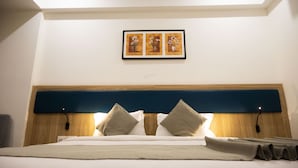 A neatly made bed with a wooden headboard, two bedside lamps, and a framed floral artwork above it. - RAAJ SVASTIK INN (Jamnagar)