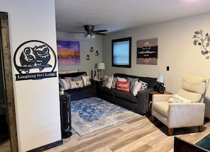 A living room with a dark grey sofa, a light grey armchair, a blue patterned rug, a ceiling fan, and wall art including an owl logo and a mountain landscape.
