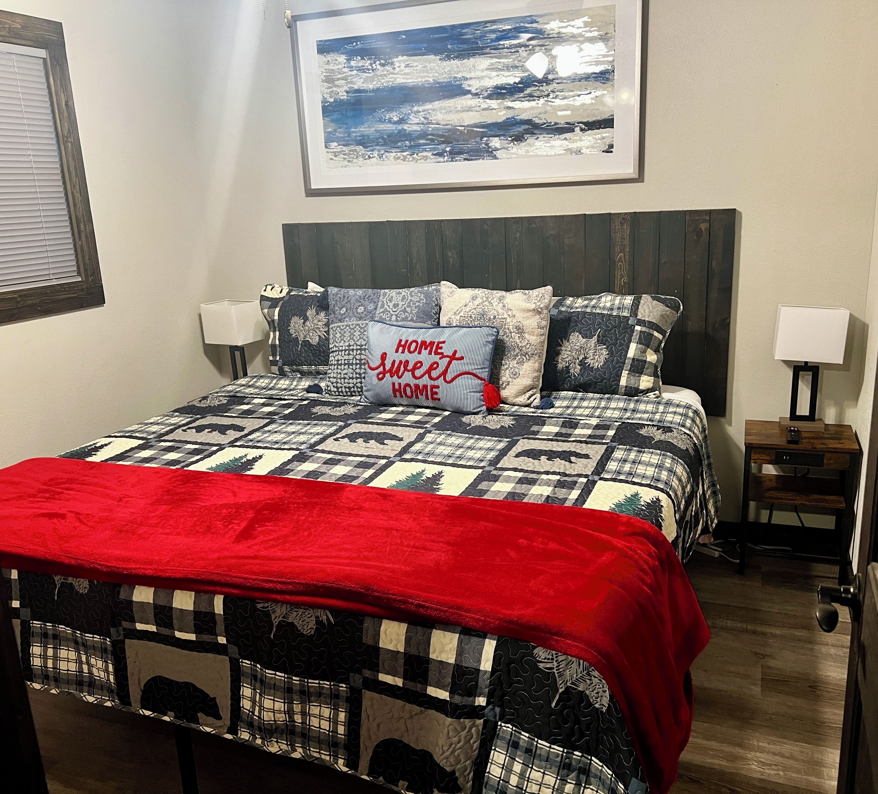 A bedroom with a bed, pillows, a red throw blanket, a headboard, a nightstand, a lamp, and a framed picture on the wall.