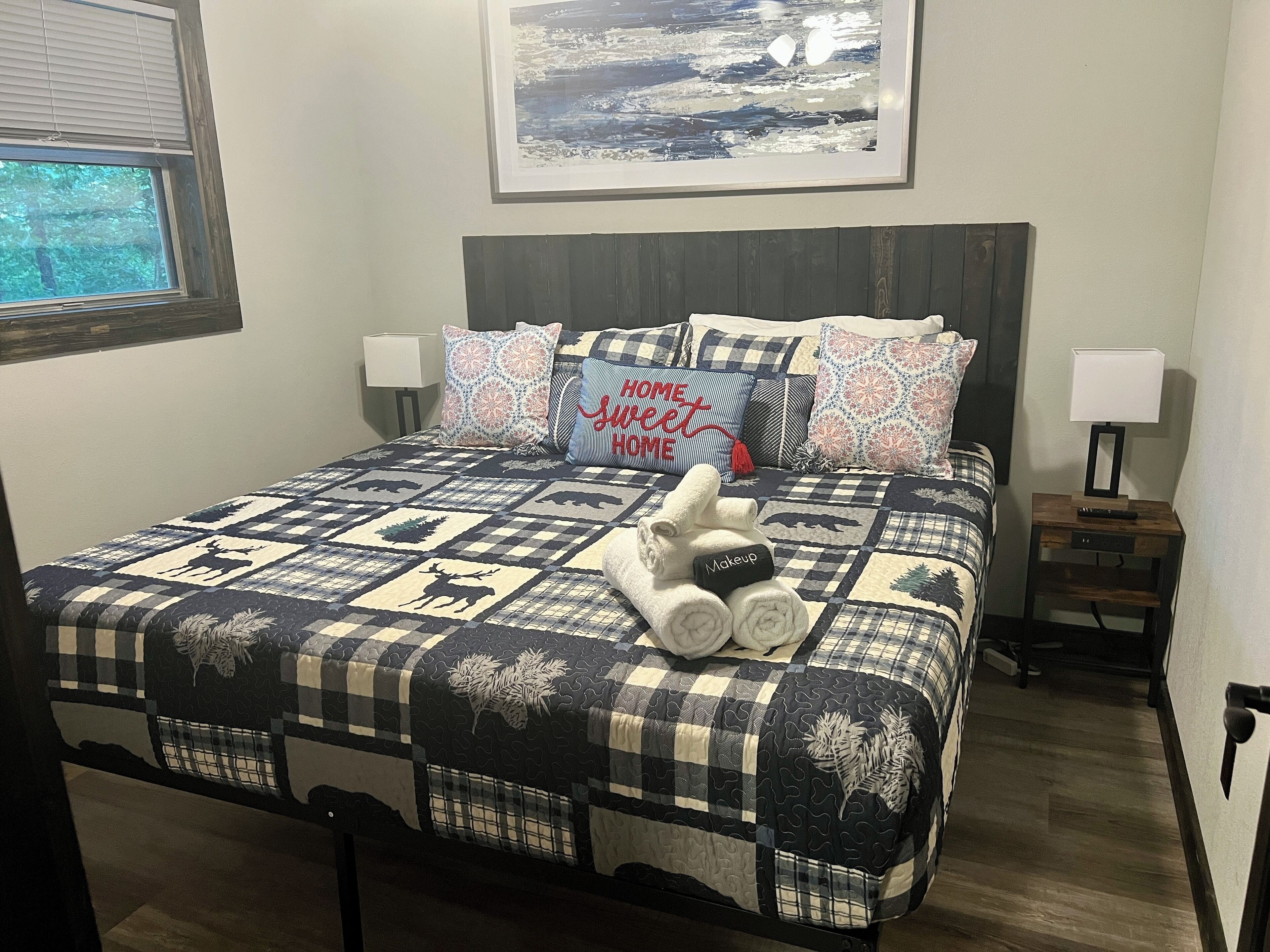 A neatly made bed with a quilt featuring animal prints, multiple pillows with various patterns, and a bedside table with a lamp and remote control.