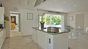 A modern kitchen with a central island, a dining area with clear chairs, and a view of greenery outside.