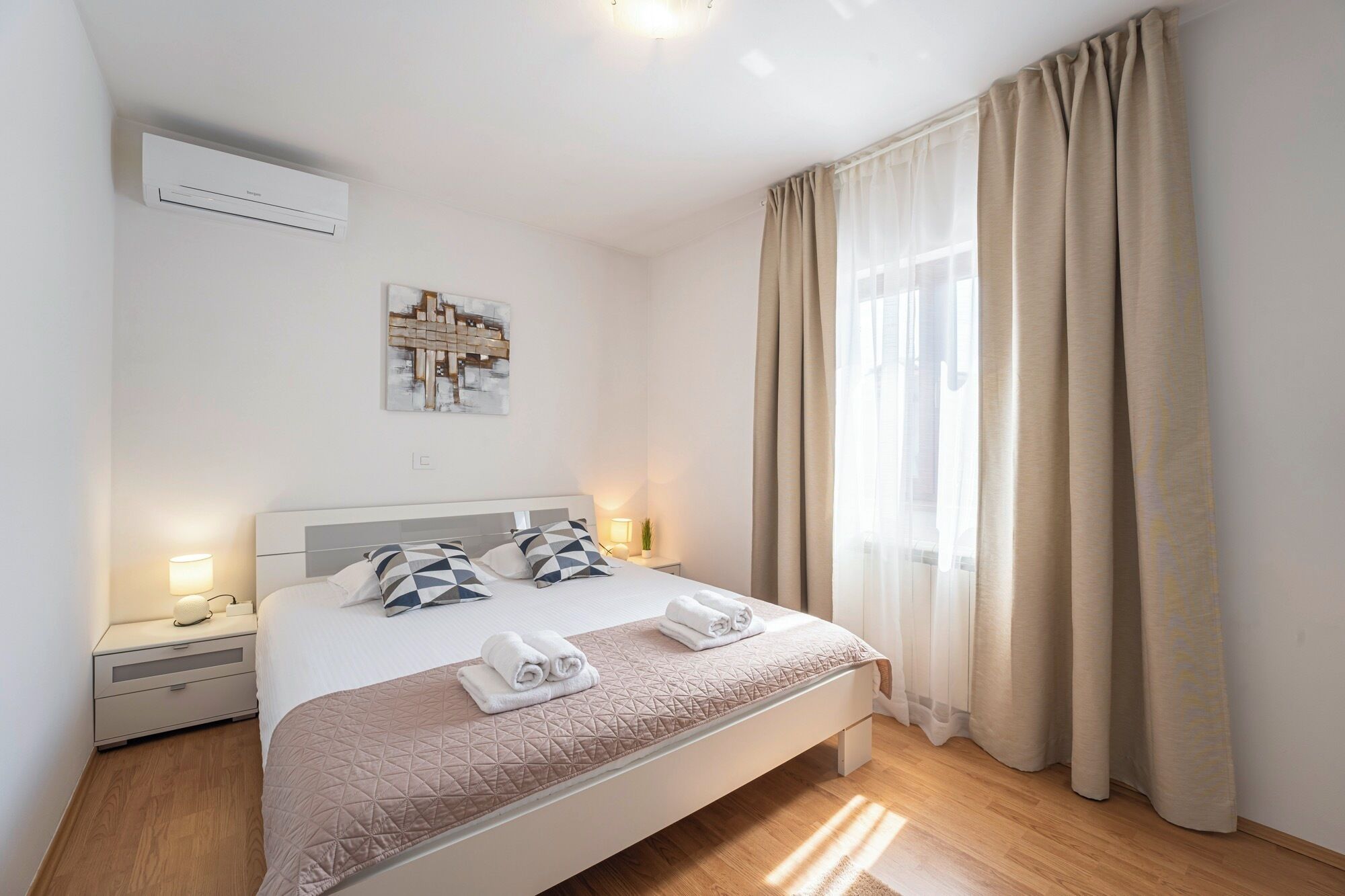 Une chambre d’hôtel avec un lit, des lampes de chevet, un climatiseur fixé au mur, une fenêtre avec des rideaux et une œuvre d’art abstraite.