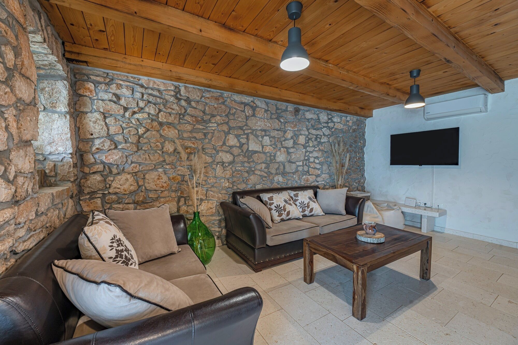 Un salon chaleureux avec des murs en pierre, des poutres apparentes en bois au plafond, un canapé en cuir, une table basse et un téléviseur à écran plat fixé au mur.