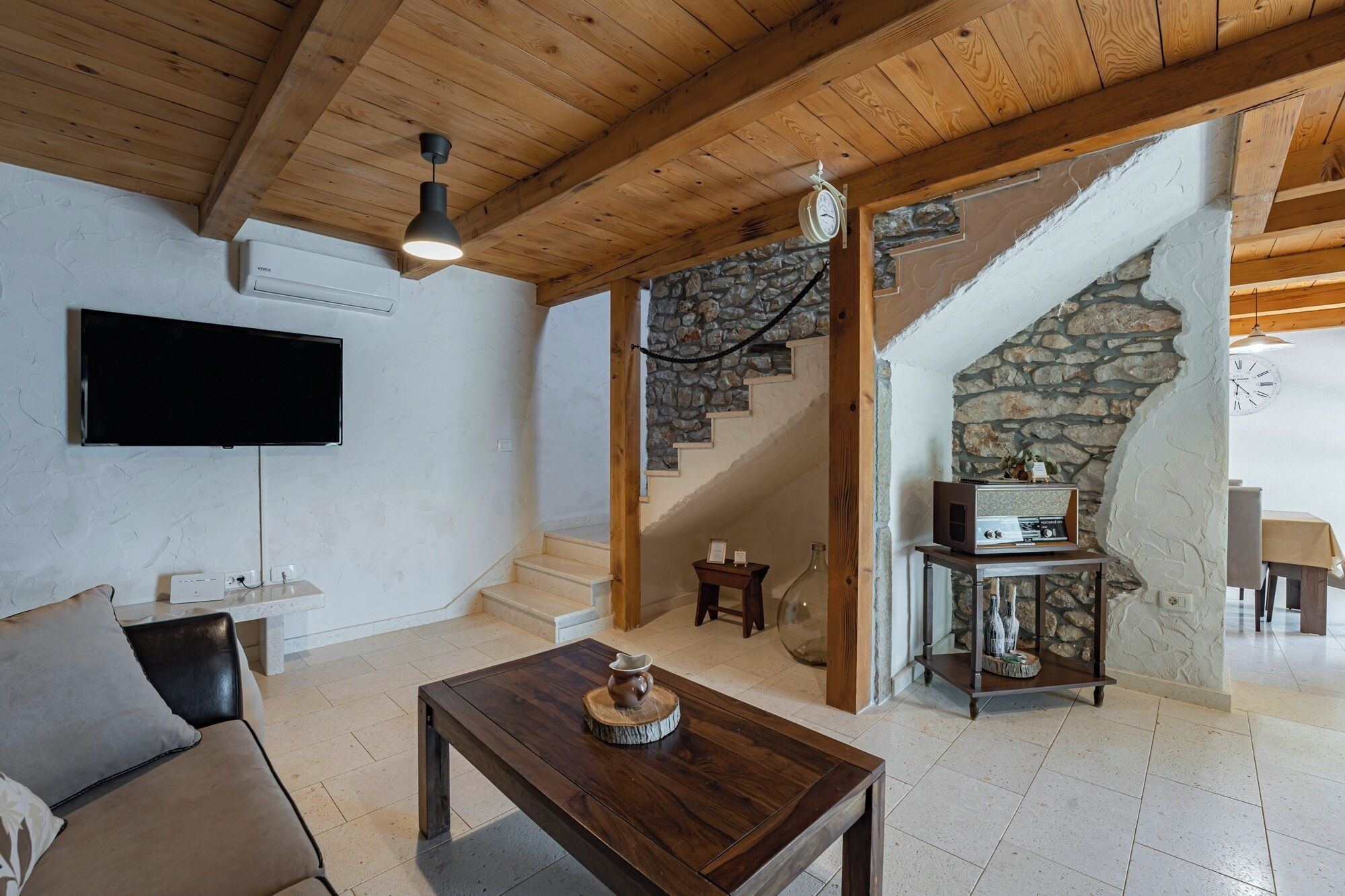 A living room with a wooden ceiling, a stone staircase, a flat-screen TV, a coffee table, and a small table with a vintage radio.