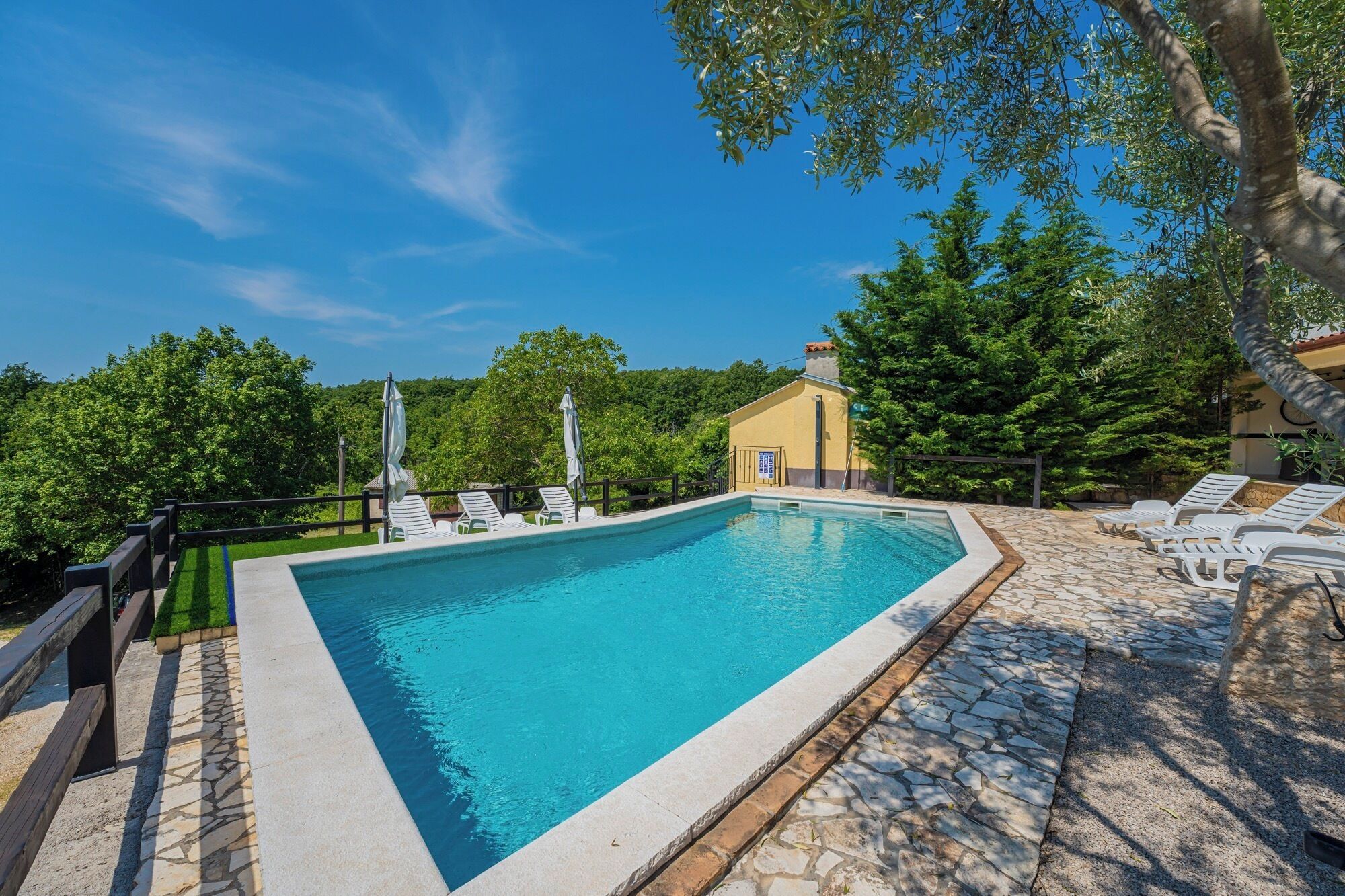 A rectangular swimming pool with a stone border, surrounded by lounge chairs and umbrellas, set against a backdrop of trees and a clear sky.