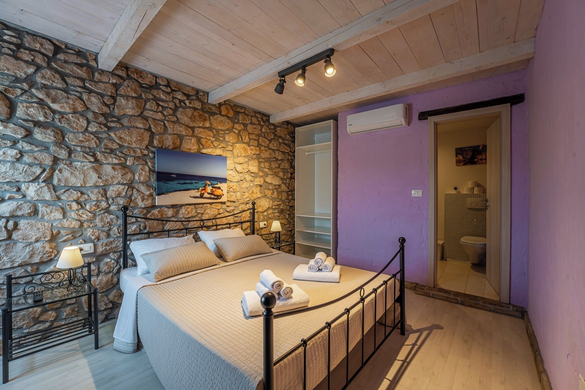 A bedroom with a stone wall, a bed with pillows, a nightstand with a lamp, and a painting of a beach scene.