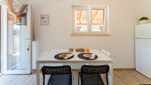Een eethoek met een tafel voor twee personen, twee stoelen, een raam met houten luiken, een witte koelkast en een deur met een ruit.