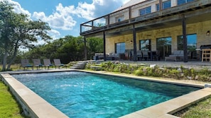 A modern outdoor pool area with lounge chairs, a covered patio, and a stone wall. - The Point-Hill Country Views-Pool+Hot Tub (Fredericksburg)