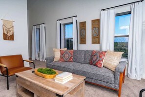 A living room with a grey sofa, a wooden coffee table, and a chair. There are two windows with curtains, a macrame wall hanging, and a view of a blue tent. - The Point-Hill Country Views-Pool+Hot Tub (Fredericksburg)