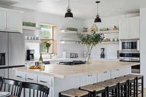 A modern kitchen with white cabinetry, a large island, bar stools, and pendant lighting. - The Point-Hill Country Views-Pool+Hot Tub (Fredericksburg)