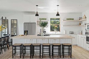 A spacious kitchen with a large island, white cabinetry, and black bar stools. - The Point-Hill Country Views-Pool+Hot Tub (Fredericksburg)