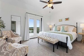A bedroom with a bed, a bench, a ceiling fan, a window, and a door. - The Point-Hill Country Views-Pool+Hot Tub (Fredericksburg)