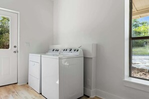 A laundry room with a washer and dryer, a door, and a window with a view of greenery. - The Point-Hill Country Views-Pool+Hot Tub (Fredericksburg)