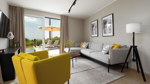 En moderne stue med sofa, gul lænestol og sofabord. Der er et stort vindue med gardiner, en gulvlampe og et indrammet billede på væggen.