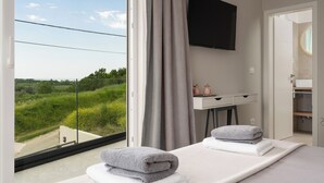 Et moderne hotelværelse med et stort vindue, der giver udsigt over grønne områder og et fladt landskab. Der er en seng med håndklæder, et skrivebord med et fladskærms-tv og et lille bord med pynt.