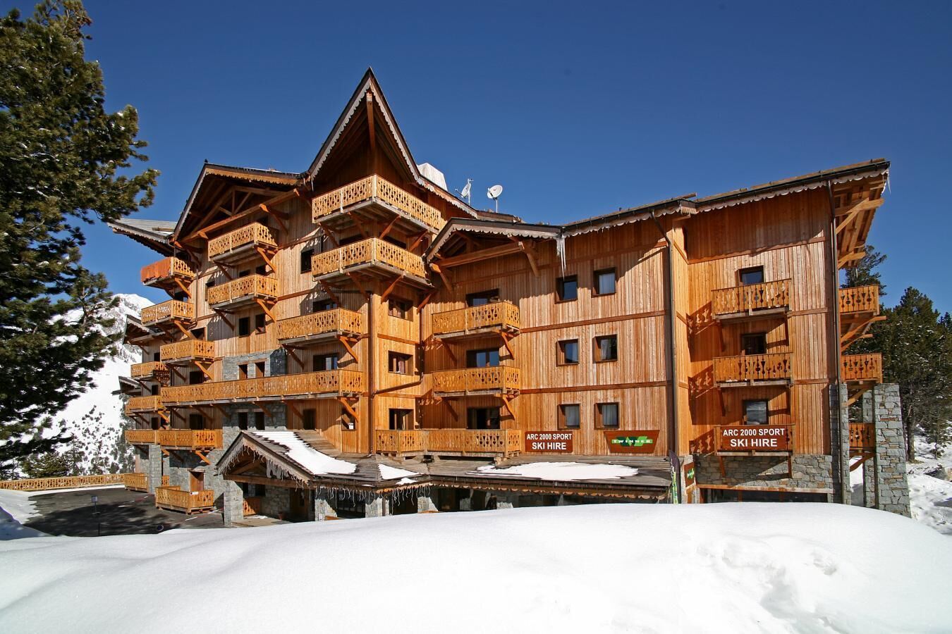 A multi-story wooden building with balconies, ski hire signs, and a snowy landscape.