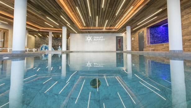 Une piscine intérieure spacieuse au design moderne, avec un plafond en bois et une enseigne indiquant « CHALET ALTITUDE ».