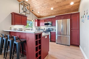 A kitchen with red cabinets, stainless steel appliances, a wooden ceiling, and a bar area with stools.