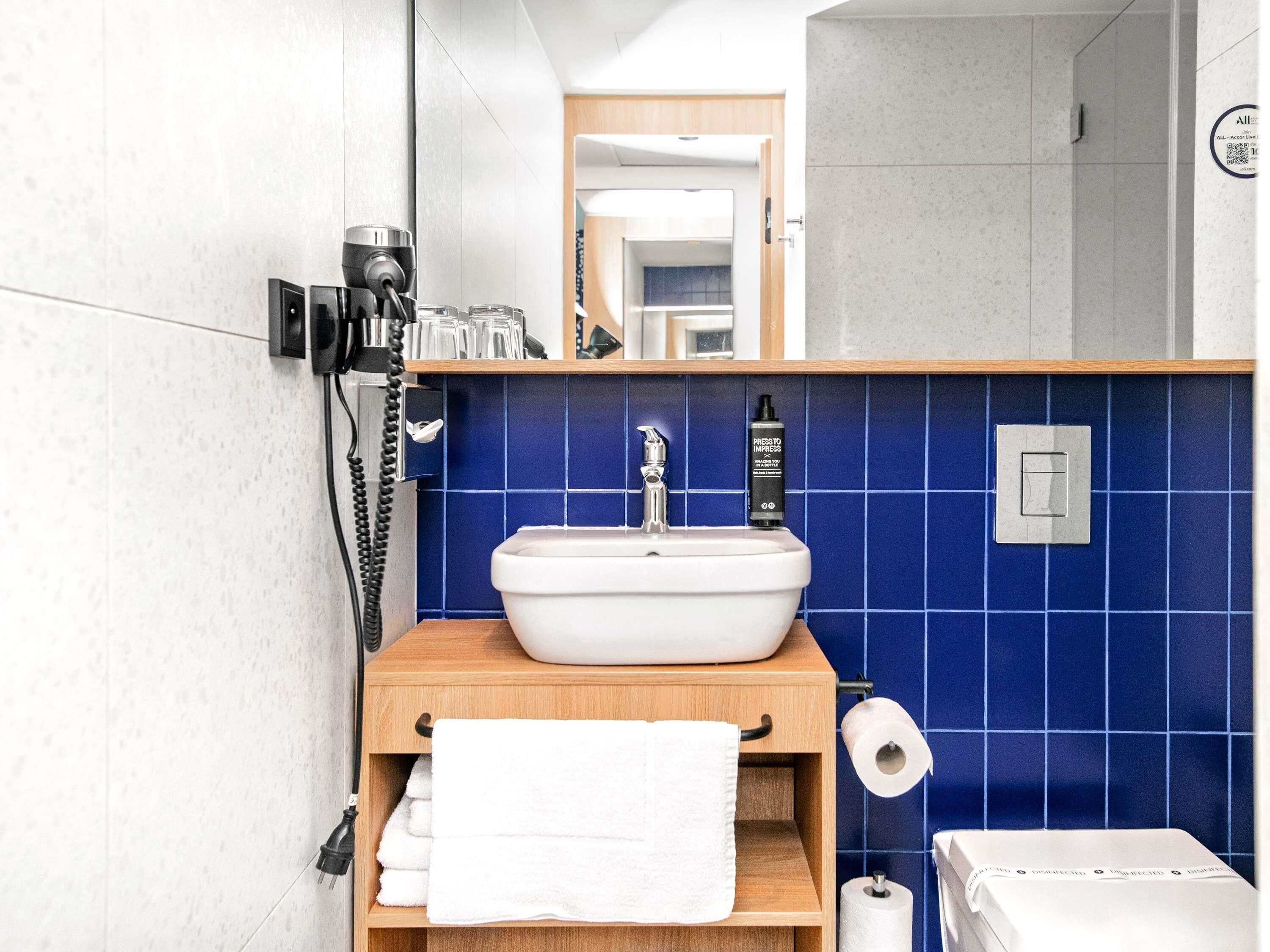 A modern bathroom with a white sink, blue tiles, a wooden vanity, a towel rack, and a visible phone.