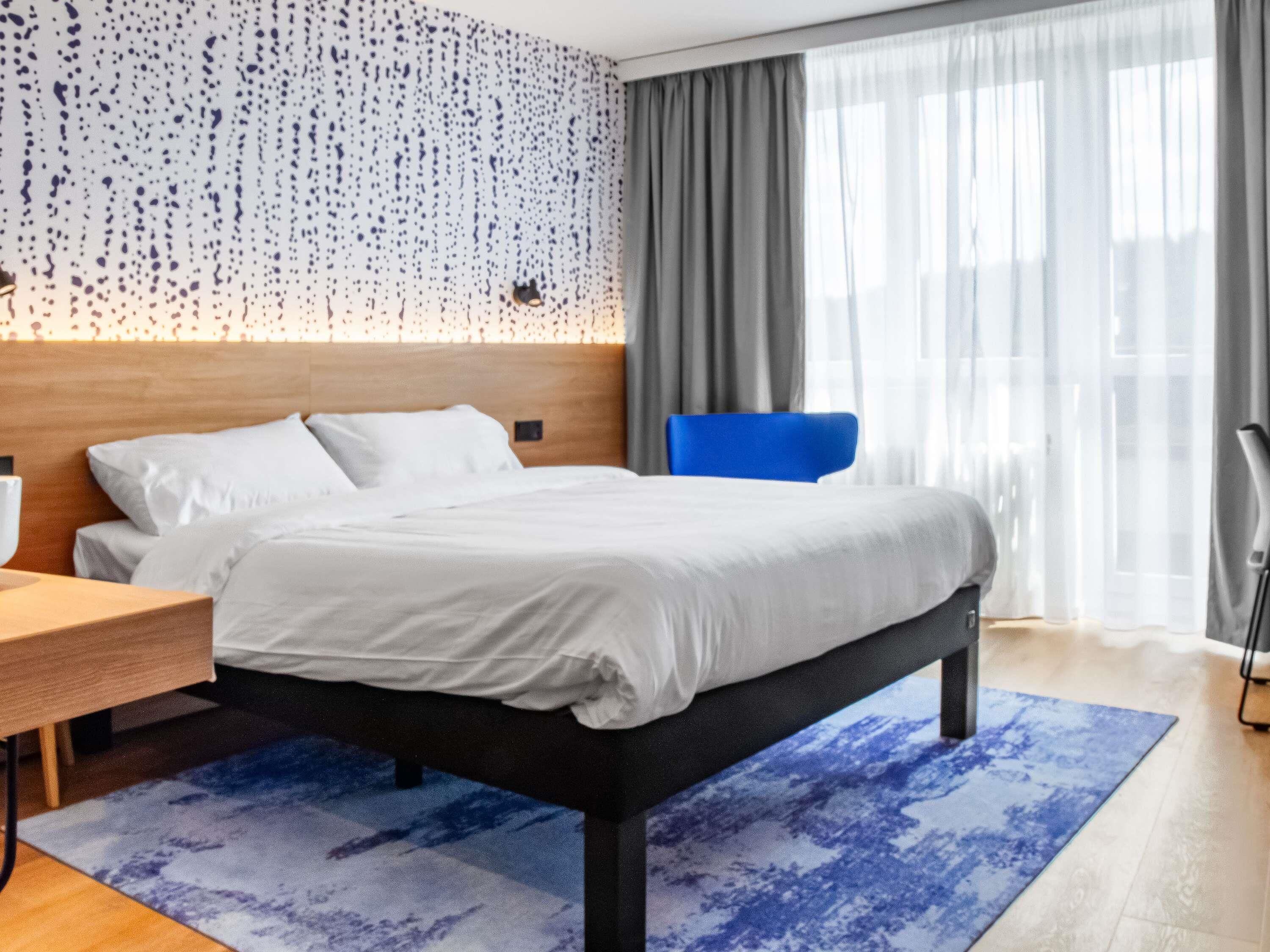 A modern hotel room with a large bed, a wooden headboard, a desk, and a chair.